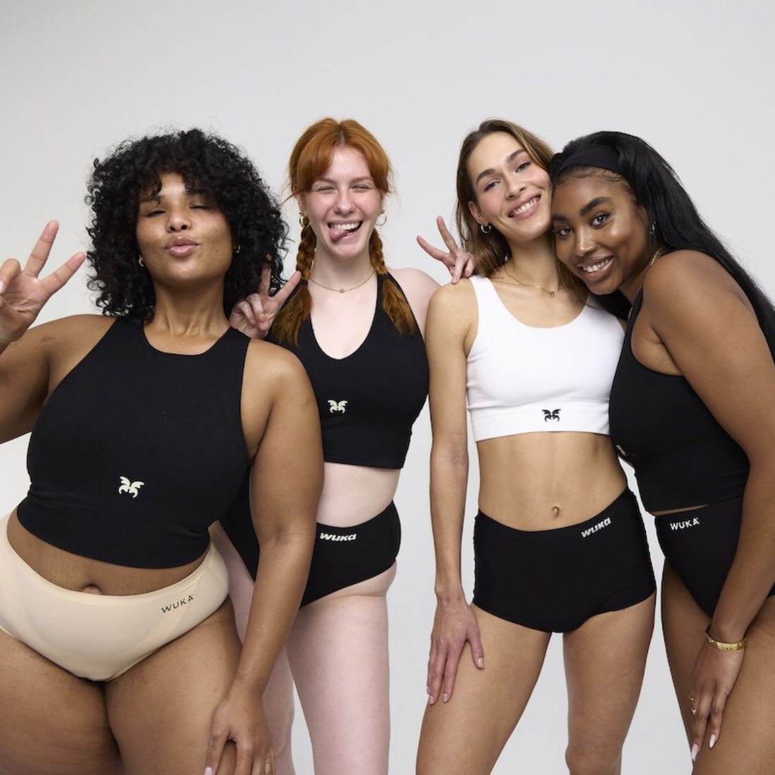 Four young women wearing WUKA period pants and crop tops pose confidently, smiling and flashing peace signs - celebrating body positivity and sisterhood.